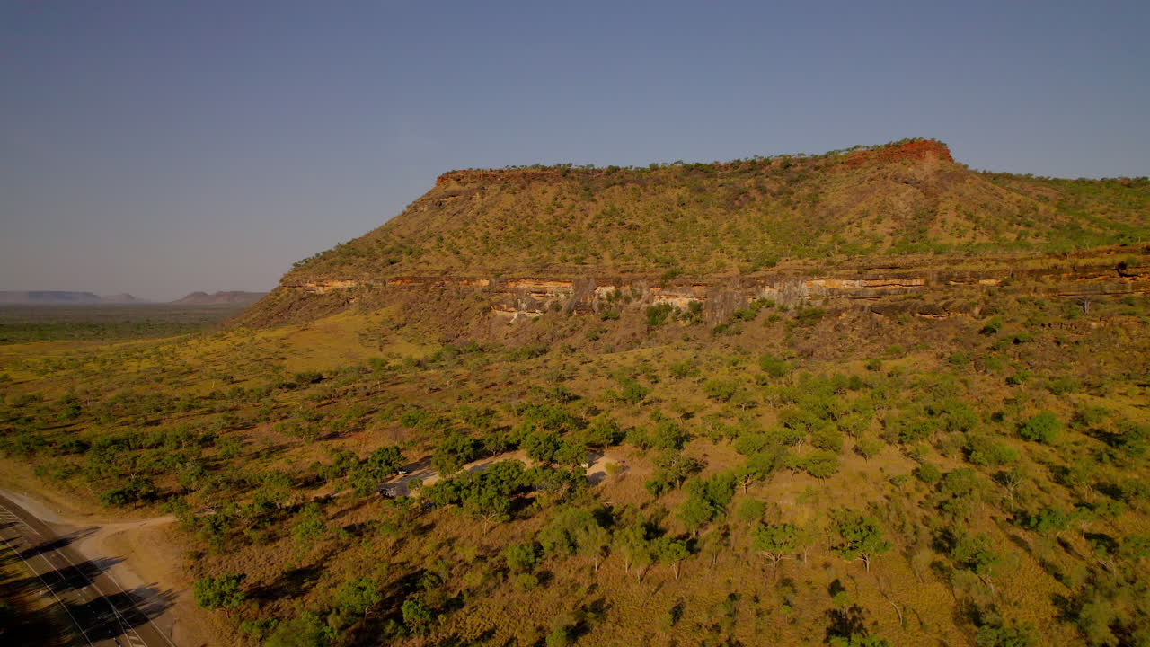 Ripresa cinematografica con drone che vola all'indietro sulla Saddle Creek Rest Area nel Territorio del Nord Australia durante la calda luce serale, rivelando scogliere aspre, boscaglia e un'atmosfera da viaggio on the road nel remoto entroterra.
