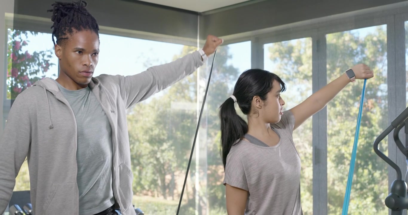 Diverse pair in sportswear with resistance bands, man guiding raises, woman following at home gym