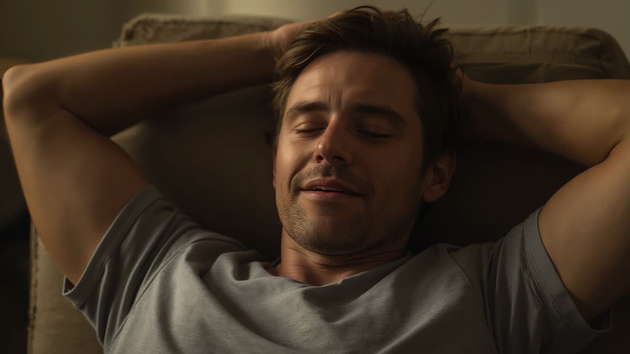 Leaning back, man in gray T-shirt holding hands behind head on home armchair, closing eyes, resting