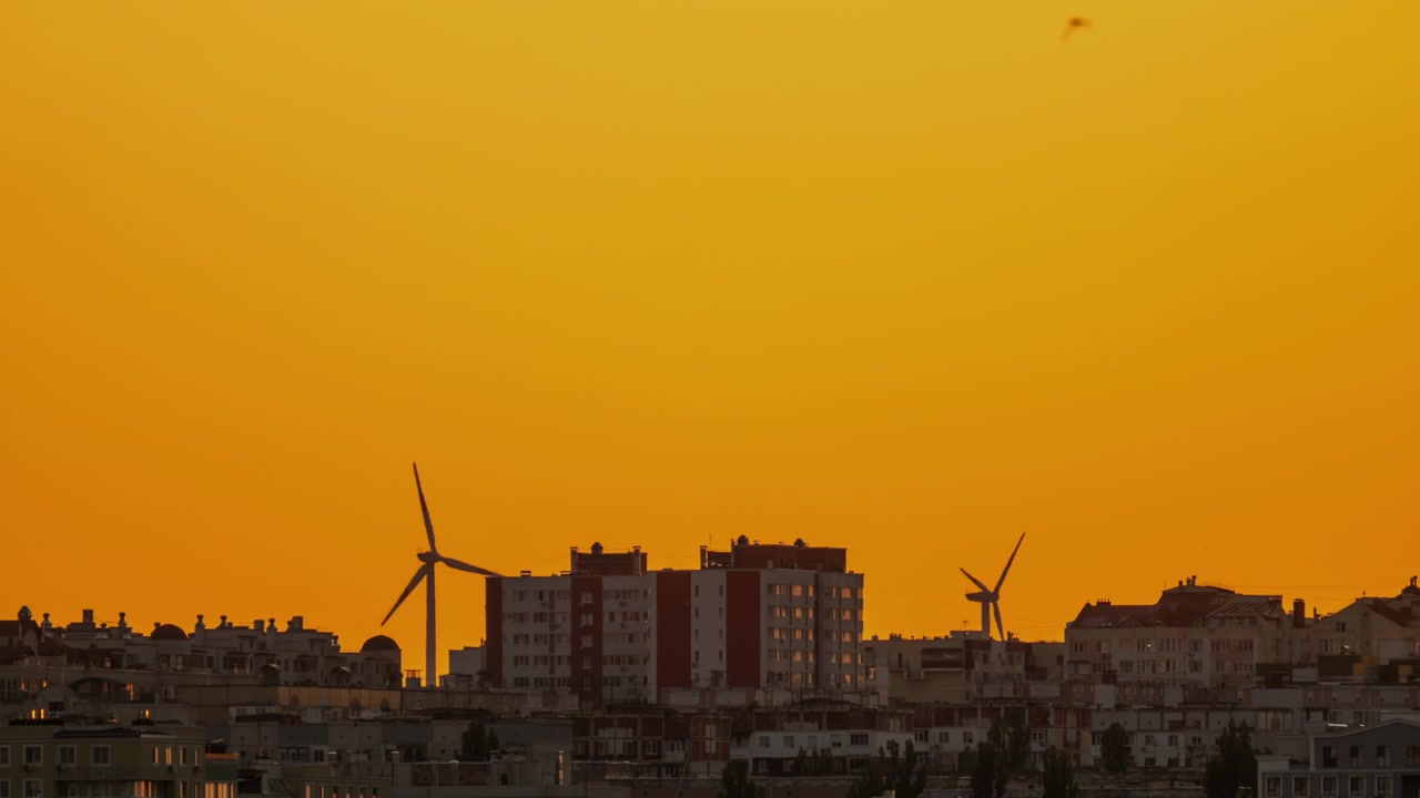 Lapso de tiempo de aerogeneradores silueteados girando sobre el horizonte de una ciudad durante un atardecer dorado
