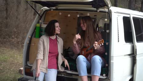 two young girl friends chat in the back of a caravan in the middle of the forest. one of them plays the ukelele.