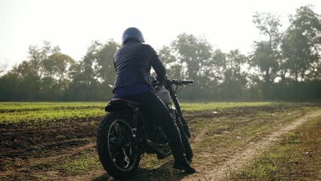 Rückansicht-Eines-Mannes-Mit-Helm-Und-Lederjacke,-Der-Auf-Sein-Fahrrad-Zugeht-Und-Den-Motor-Startet,-Während-Er-Im-Gelände-Steht