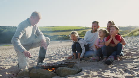 multi-generation family having evening barbecue around fire on beach vacation