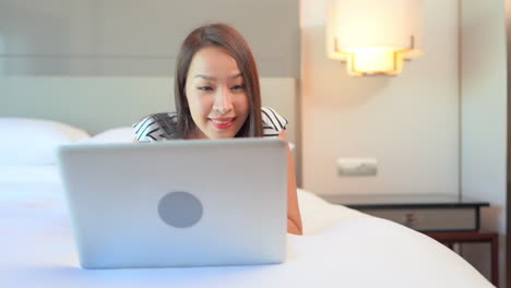 close-up of a young woman lying on a bed working happily on her laptop