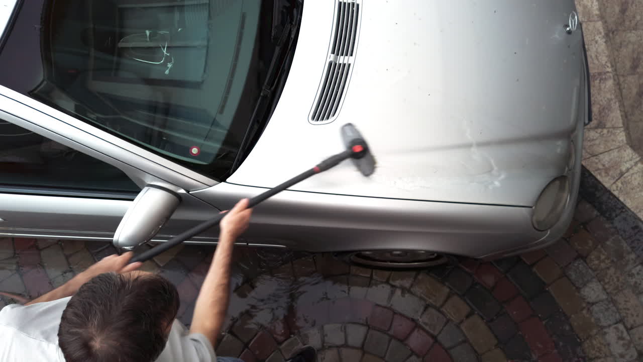 A person is washing a silver car in a driveway. The focus is on the methods used, showing care and attention to detail while cleaning the vehicle's surface