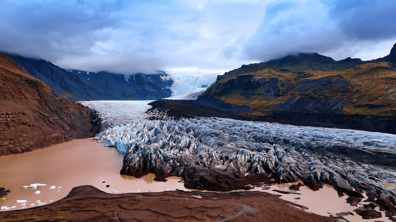 Ancient picturesque glacier descending from the mountain. Ice boulders go down the lake. Thick cloudscape covers the rocks peaks.