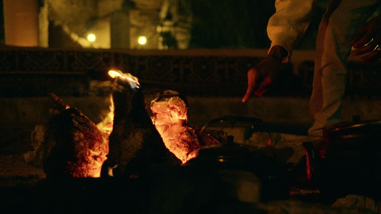 Cena noturna cinematográfica de uma pessoa fervendo um bule de ferro grande sobre uma lareira ao ar livre. As toras queimando brilham enquanto as mãos ajustam a fogueira, criando uma atmosfera aconchegante na escuridão