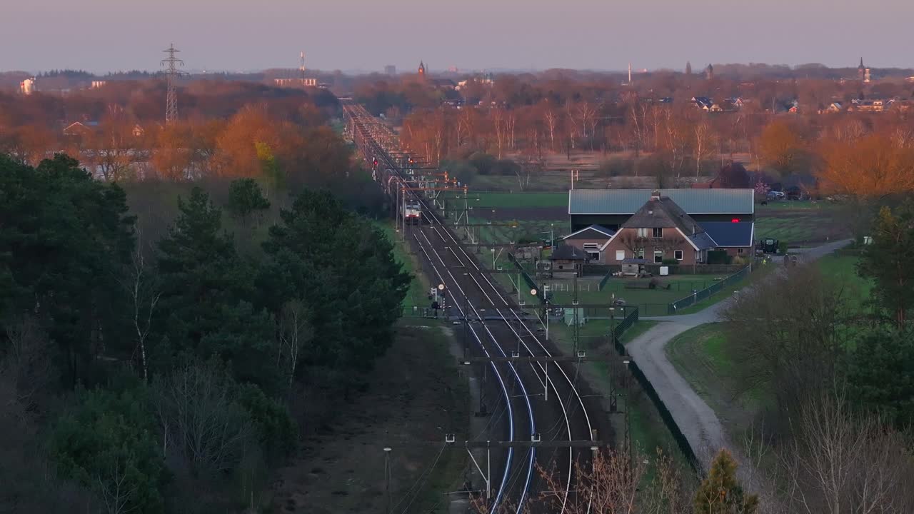 Drone flies over railway at dusk in the Netherlands, capturing a train, a house, and a distant town in soft evening light.