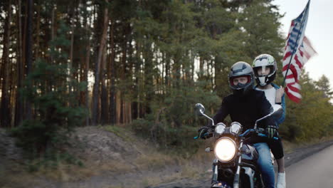 a pair of bikers travels on a motorcycle followed by an american flag