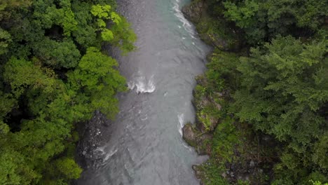 Drone-shot-of-clear-shallow-turquoise-river-in-the-middle-of-a-forest-on-a-cloudy,-moody-day