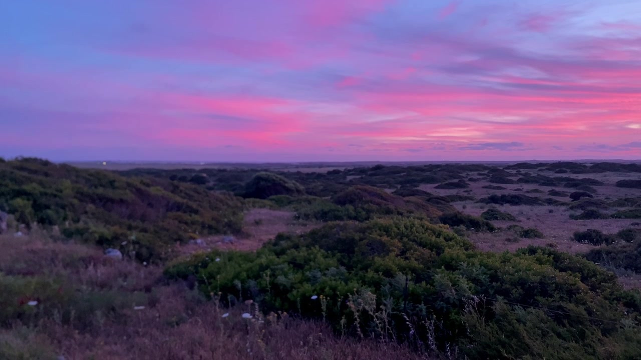 Kystlandskap med lav middelhavsvegetasjon, med en levende rosa og lilla solnedgang som skaper en dramatisk himmel.