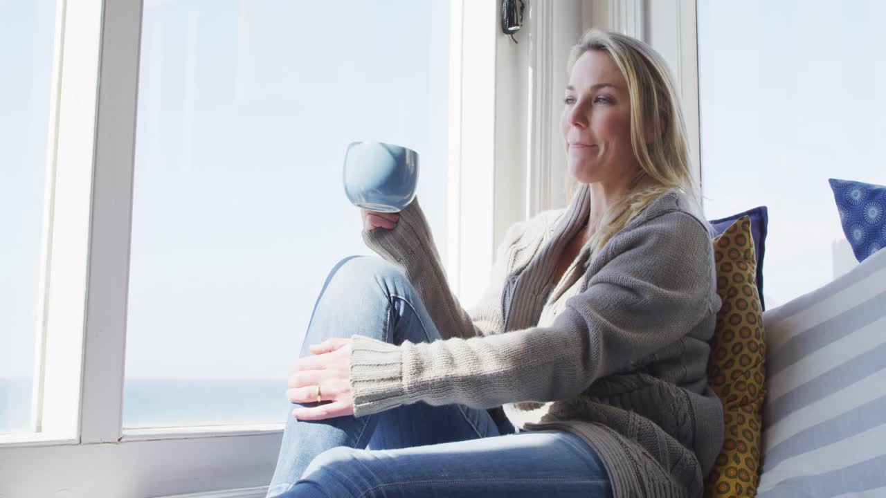 Kaukasisk moden kvinde kigger ud ad vinduet og drikker kaffe.