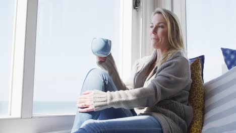 Caucasian-mature-woman-looking-through-the-window-and-drinking-coffee