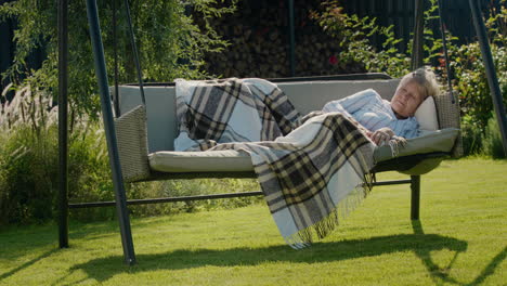 an elderly woman sleeps in a garden swing in the backyard of a house