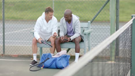 vielfältige glückliche männliche freunde, die auf einem smartphone sitzen, sitzen auf einer bank auf einem tennisplatz, slow motion