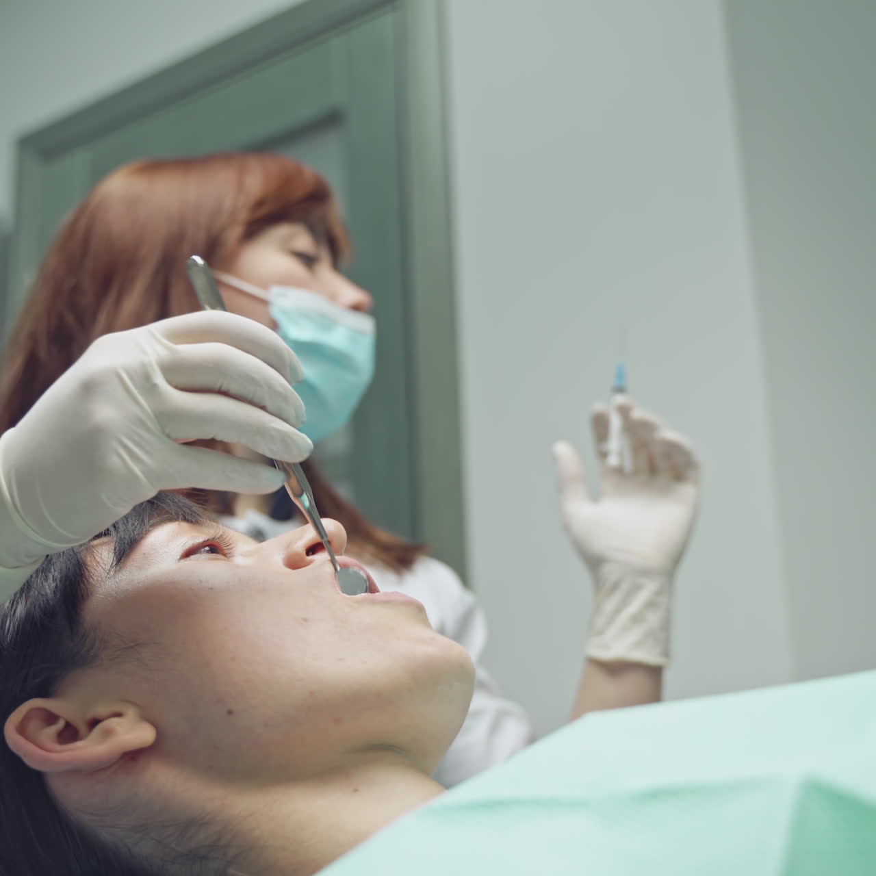 Female stomatologist applying anesthetic injection before oral surgery operation in clinic. Pretty dentist using mouth mirror and medicine syringe during her work. Close-up