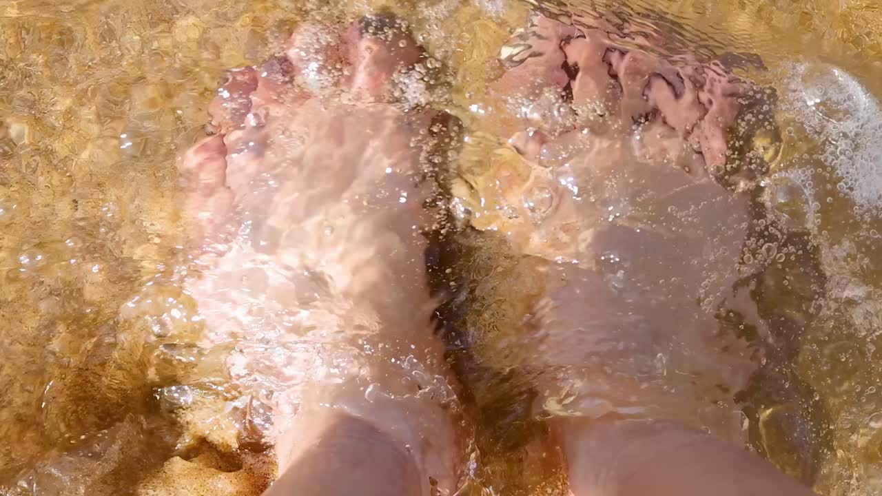 Close-up of feet submerged in clear, sunlit waves on a sandy beach.