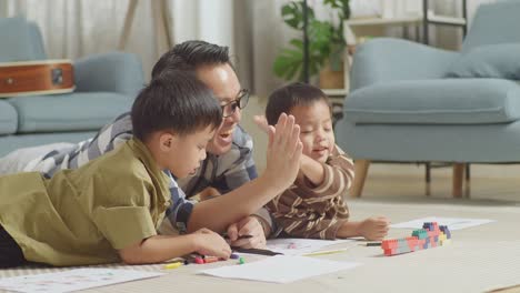 close up of asian father and sons on the floor in the room with plastic toy brick giving high five celebrating on success drawing together at home