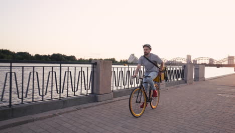 food delivery rider guy wearing thermal backpack rides a bike next to a river to deliver orders for clients and customers