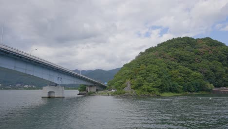 Navigating-under-a-concrete-bridge-on-a-Japanese-lake-and-surrounded-by-green-hills,-the-boat's-view-unfolds-beneath-the-overcast-sky,-creating-a-serene-atmosphere