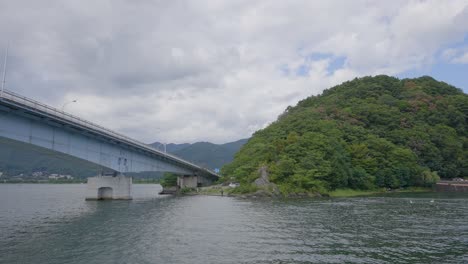Navigating under a concrete bridge on a Japanese lake and surrounded by green hills, the boat&#039;s view unfolds beneath the overcast sky, creating a serene atmosphere