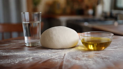 Dough, water, and olive oil on a floured wooden surface in a kitchen