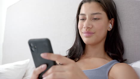 adolescente biracial feliz sentada en la cama por la mañana usando teléfono inteligente y auriculares, cámara lenta