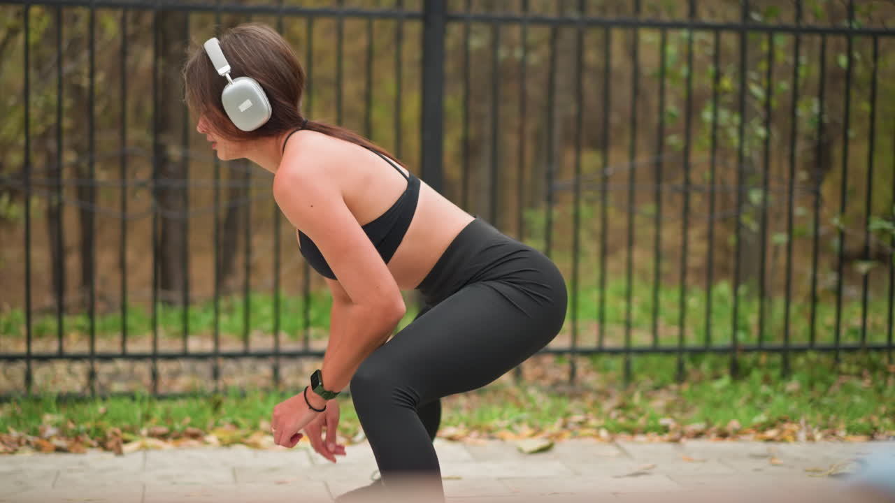 Lady in black workout gear stretching her legs outdoors near iron fence while listening to music with headphones, she is focusing on lap exercises in a park setting