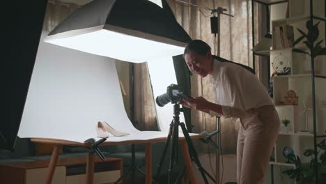 asian female photographer comparing photos on a smartphone to camera while taking photos of women's shoes in home studio