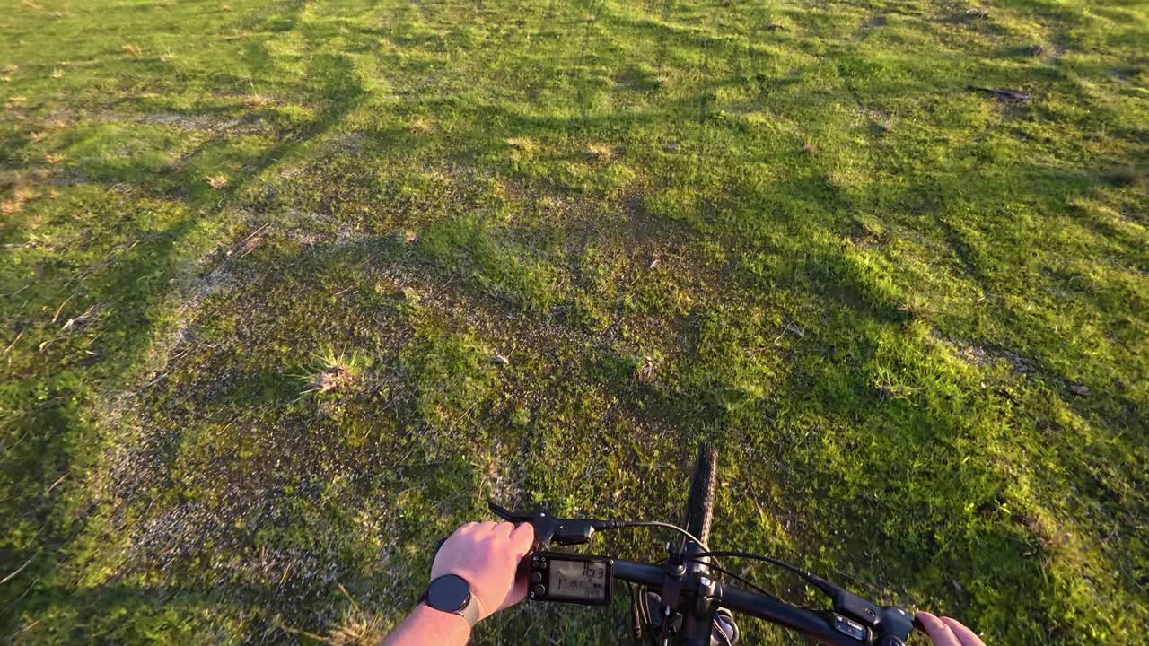 초록색 잔디을 가로질러 자전거를 타고 가는 자전거 타는 사람의 포인트 오브 뷰, 손으로 바를 잡고 앞에 있는 빛이 비치는 지형.