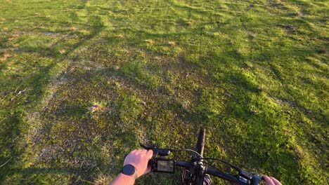 una toma de punto de vista de un ciclista montando a través de un campo de hierba verde, con las manos agarrando el manillar y el terreno iluminado por el sol delante.