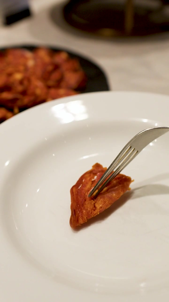 Detailní záběr ruky, která vidličkou pokládá chorizo na bílý talíř