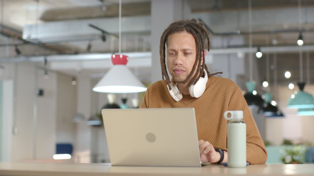 Homem afro-americano digitando no laptop no escritório, usando mouse, pegando garrafa, reagindo a um aviso na tela.