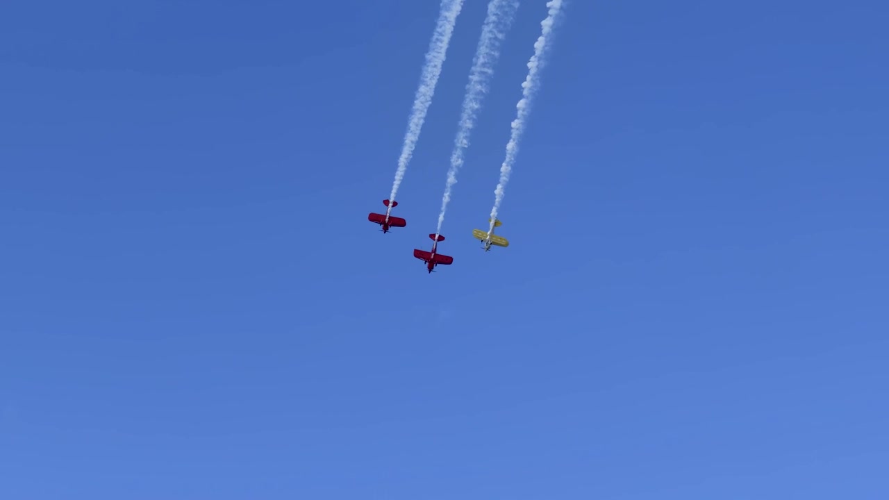 Three stunt biplanes fly in formation leaving white smoke trails against a blue sky.