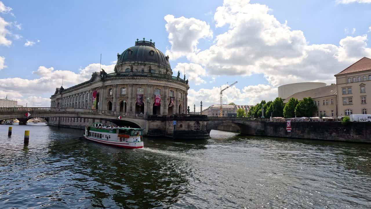 A sightseeing boat cruises along the Spree River past a historic domed museum under bright daylight, with dynamic water reflections and gentle camera pan
