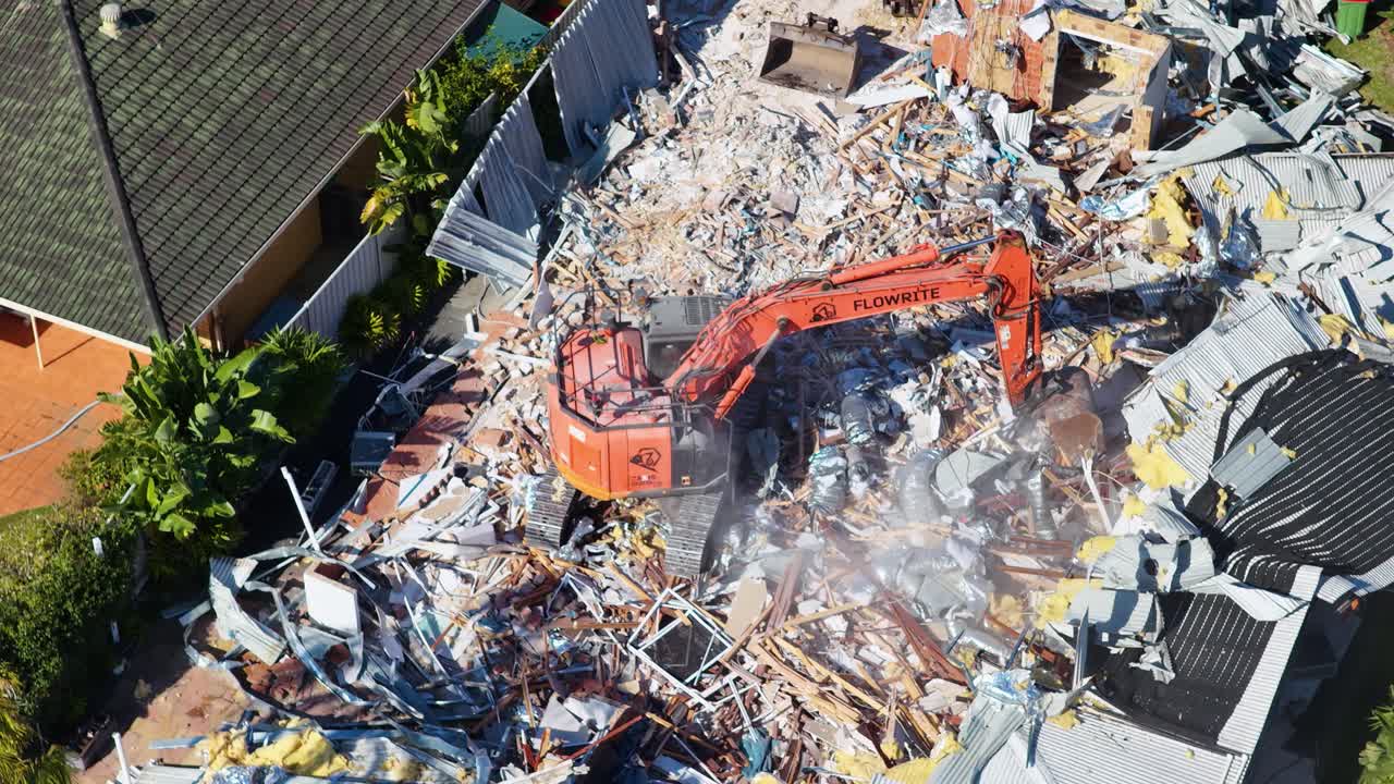 A large orange excavator actively demolishes a residential house, surrounded by rubble and dust, captured in bright daylight from a drone perspective