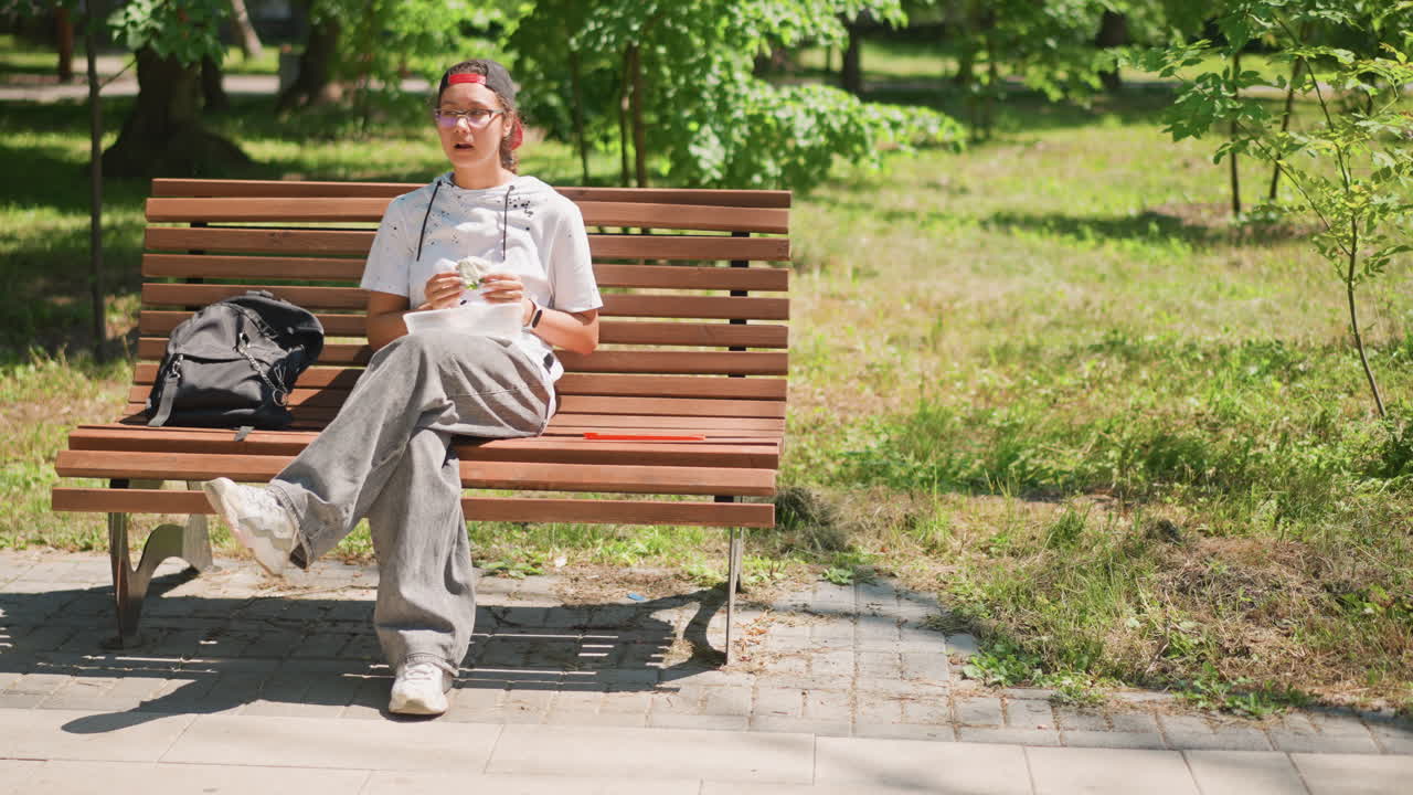 공원 벤치에 앉아 아이스크림 콘을 즐기는 백인 남성, 햇빛이 나무 사이로 쏟아지고, 옆에는 백팩이 놓여 있으며, 캐주얼 셔츠와 모자를 착용한 편안한 자세, 여름의 여유와 평화로운 도시의 녹지, 자연스러운 야외 풍경