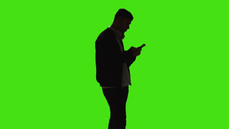 silhouette of man wearing headphones sending or reading text message or scrolling online on mobile phone standing against green screen with low key lighting