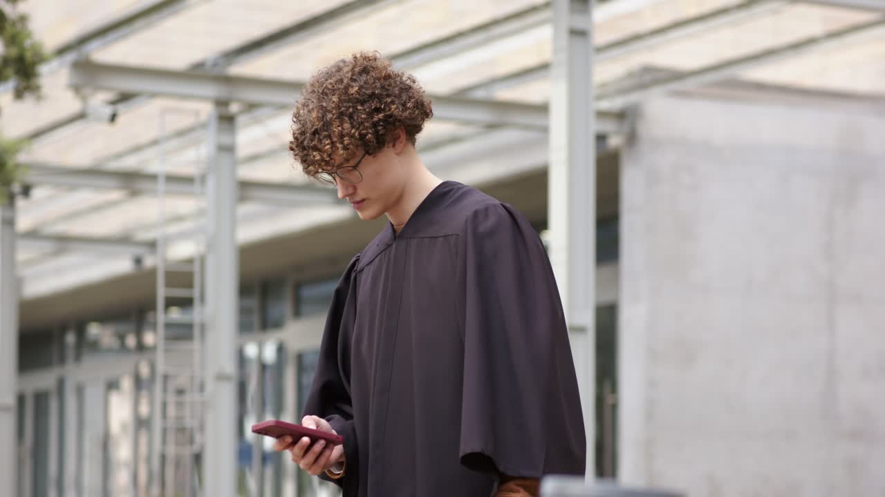 Homem de 20 e poucos anos usando beca, abrindo um estojo de certificado vermelho e olhando o celular sob o toldo após a formatura
