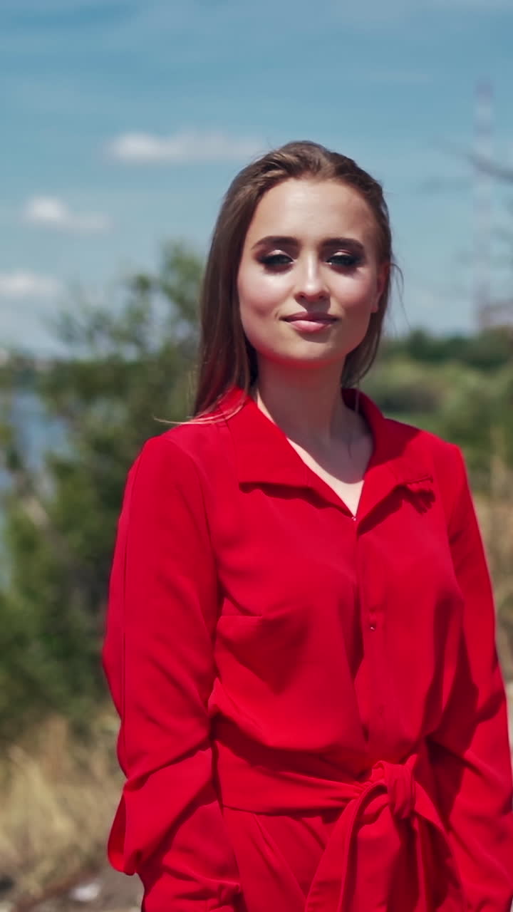 Portrait of a beautiful woman on nature background. Attractive young model in red dress with long hair standing near the river and smiling. Vertical video