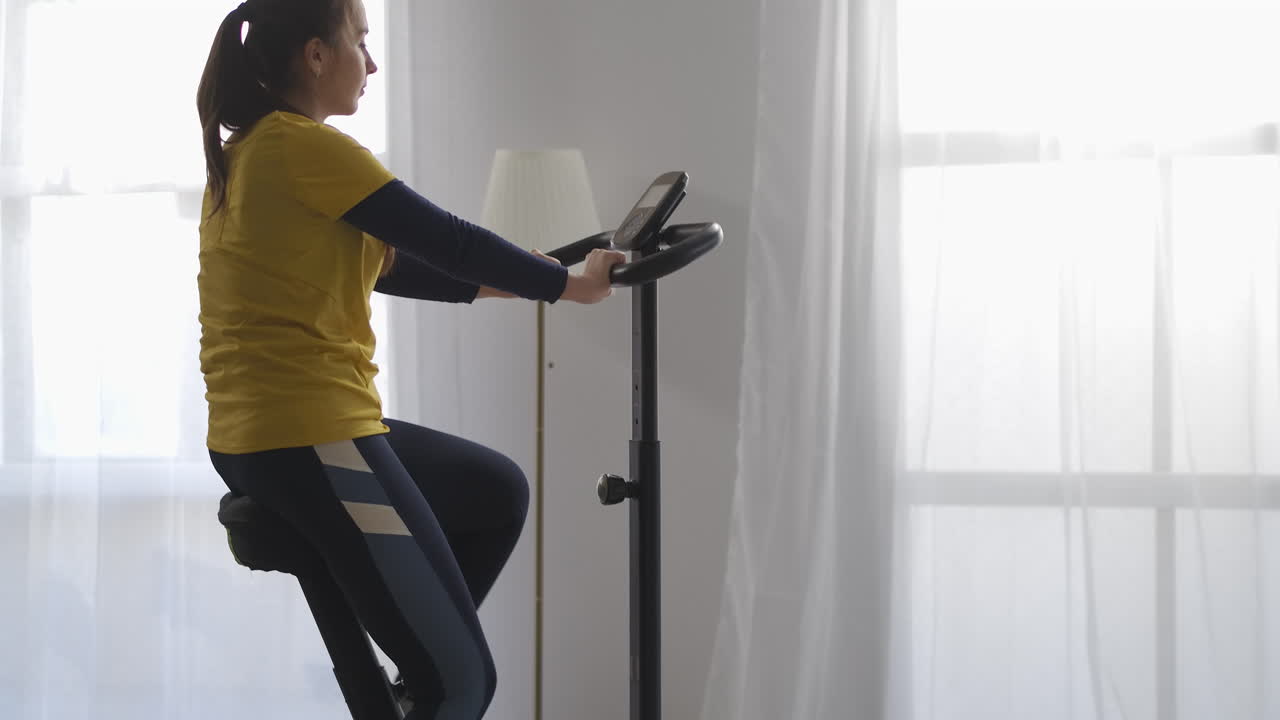 donna bruna pensierosa sta girando i pedali di una bicicletta stazionaria nel soggiorno facendo esercizio fisico per una buona figura cardio allenamento per la salute