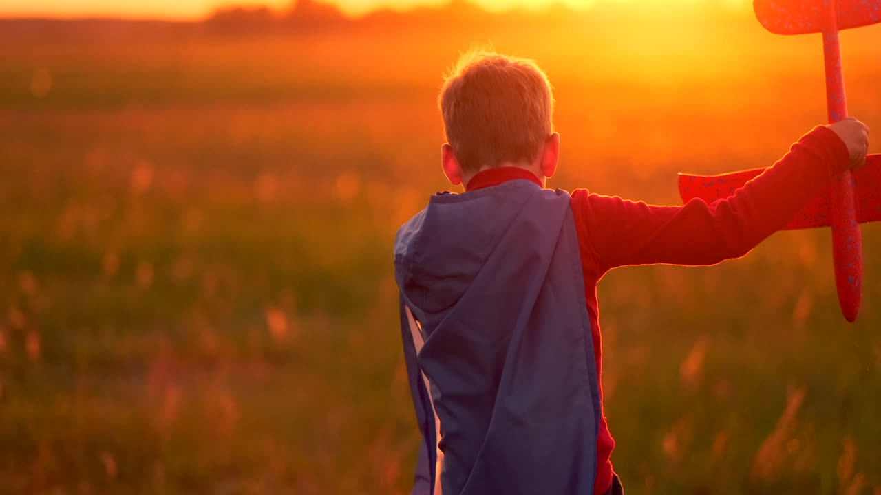 un niño corre con un impermeable rojo sosteniendo un avión riendo al atardecer en el campo de verano imaginando que es un piloto de avión jugando con un modelo de avión
