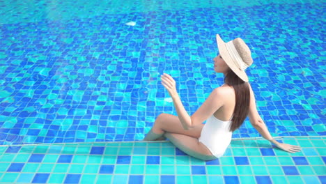 young american woman sitting on the swimming pool border with her legs inside water and touching straw sunhat brim with fingers, bora bora hotel, slow motion elevated view, copy space