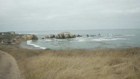 Slow-Motion-walk-on-coastal-cliff-trail-dirt-path-by-tall-dry-grass-out-to-ocean-view-and-blue-waters-on-overcast-summer-day