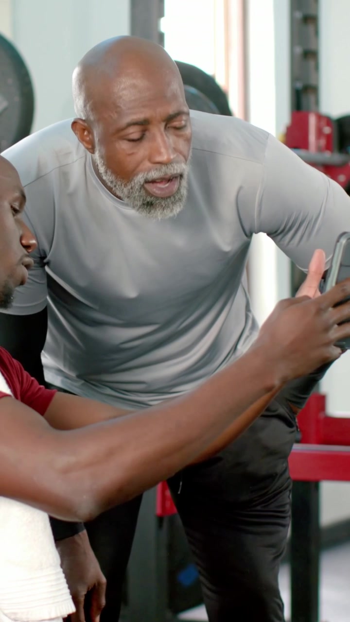 Vertical video: Leaning trainer in gray tee pointing as client tapping phone on gym bench coaching