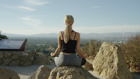 video en cámara lenta de una joven rubia vista desde atrás sentada en una roca afuera en una pose de yoga con cielo azul y hermosas vistas de fondo