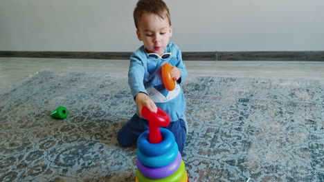 baby boy with sunglasses on his neck playing at home. caucasian toddler assembles and disassembles toy pyramid. black cat walks at backdrop.
