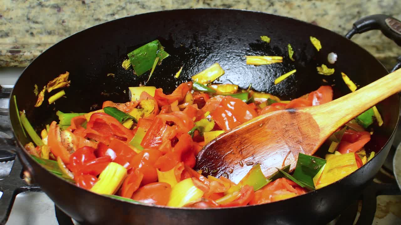 Primi piani di peperoni rossi tagliati, cipollotti e peperoncini dolci saltati in una pentola di ferro scura. Una spatola di legno è appoggiata tra le verdure colorate sul fornello della cucina.