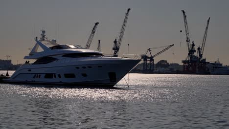 white luxury yacht is gently rocking on the calm water of a harbor, with industrial cranes looming in the background, creating a contrast between leisure and industry