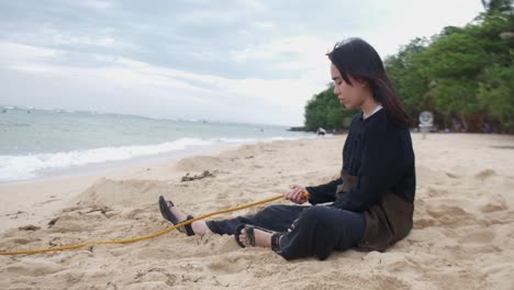 side view of pensive young woman holding rope on windy beach, symbolizing inner turmoil. -static shot